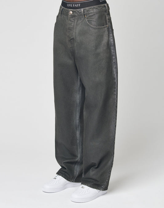 A person in LIVE FAST DIE YOUNG’s LFDY Coated Baggy Denim—baggy, wide-leg, dark grey 100% Baumwolle jeans—paired with white sneakers and LIVE FAST waistband black underwear stands against a plain white background. | model