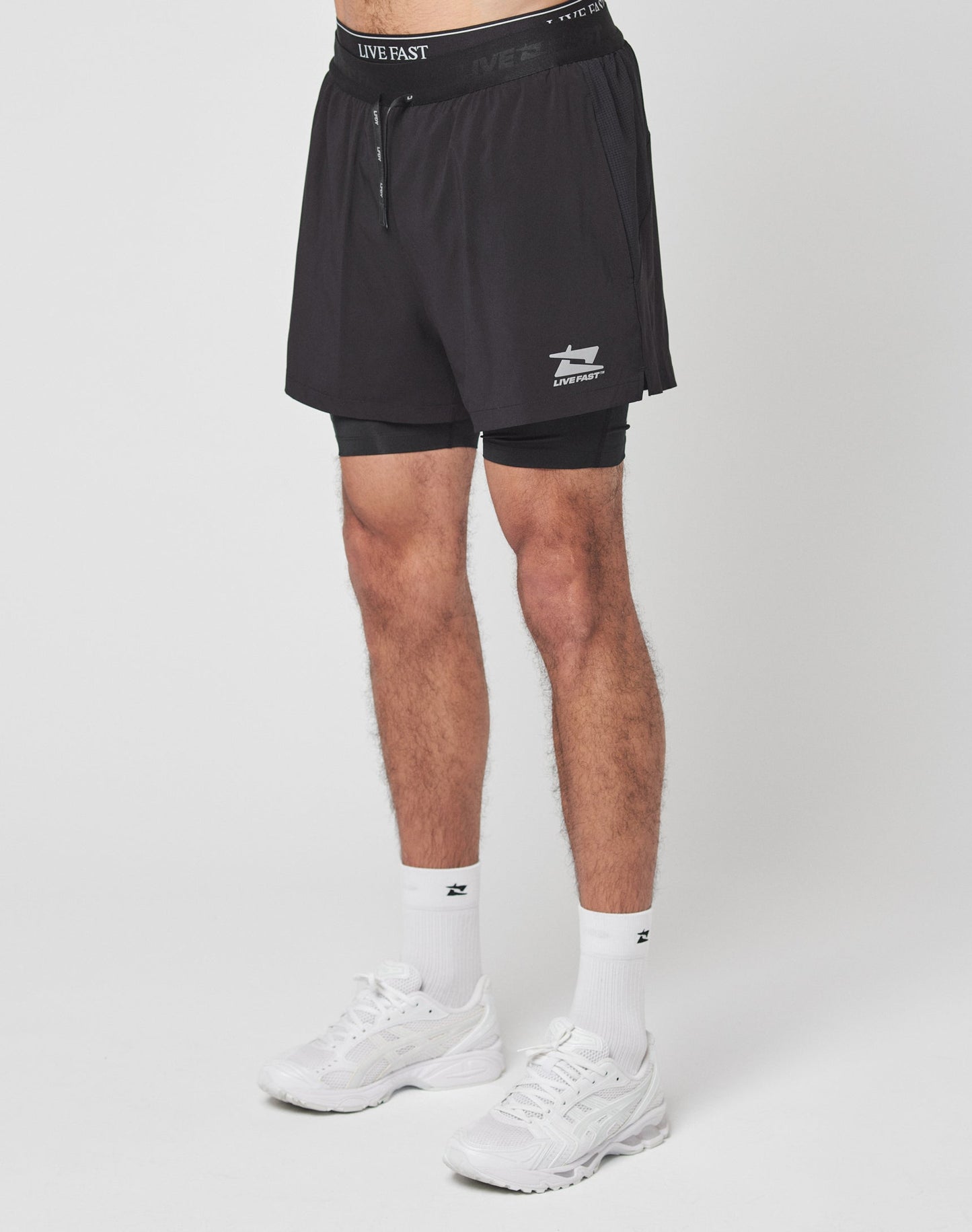 The LIVE FAST DIE YOUNG Live Fast Performance Running Shorts feature an overlapping design with built-in tights, elastic waistband with drawstrings, and a small white Live Fast Branding logo on the lower left leg, shown on a plain light gray background.| plain