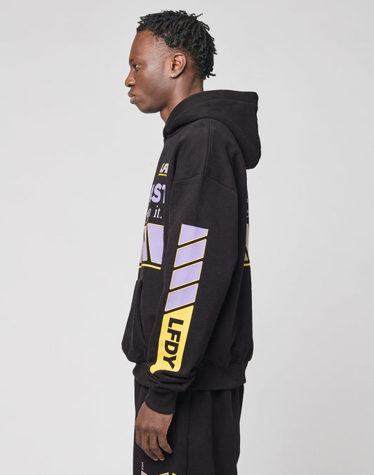 A person stands in profile against a plain light background, wearing the LFDY Racing Hooded set by LIVE FAST DIE YOUNG—an oversized black hoodie and matching pants featuring glossy yellow and purple graphics, including LFDY on the sleeve.