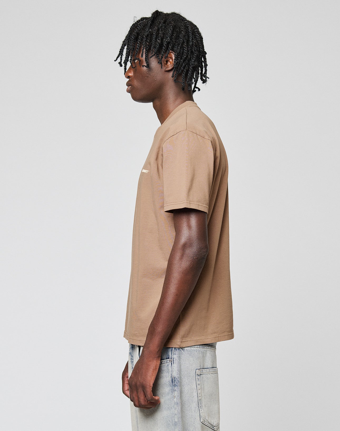 A person with short dreadlocks stands in profile, wearing the LIVE FAST DIE YOUNG Live Fast Plain Tee made from 100% Baumwolle in regular fit, paired with light-wash jeans against a plain, light gray background.