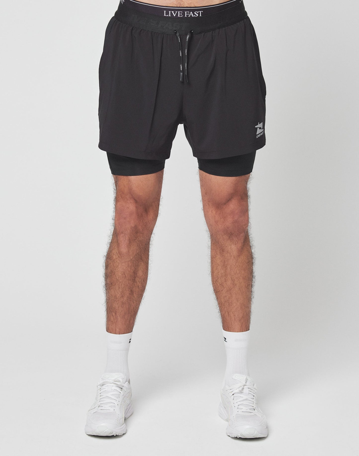 A person stands against a plain white background wearing LIVE FAST DIE YOUNG Live Fast Performance Running Shorts with integrierte Tights, a drawstring, and branded waistband, paired with white socks and sneakers.