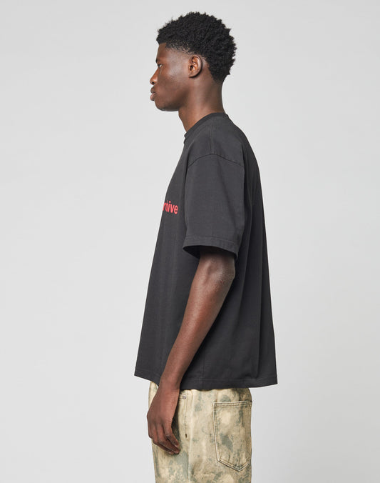 A man stands in profile against a plain background, wearing the LIVE FAST DIE YOUNG Live Fast Archive Tee—an oversize black cotton T-shirt with a red front print—paired with light, distressed jeans.