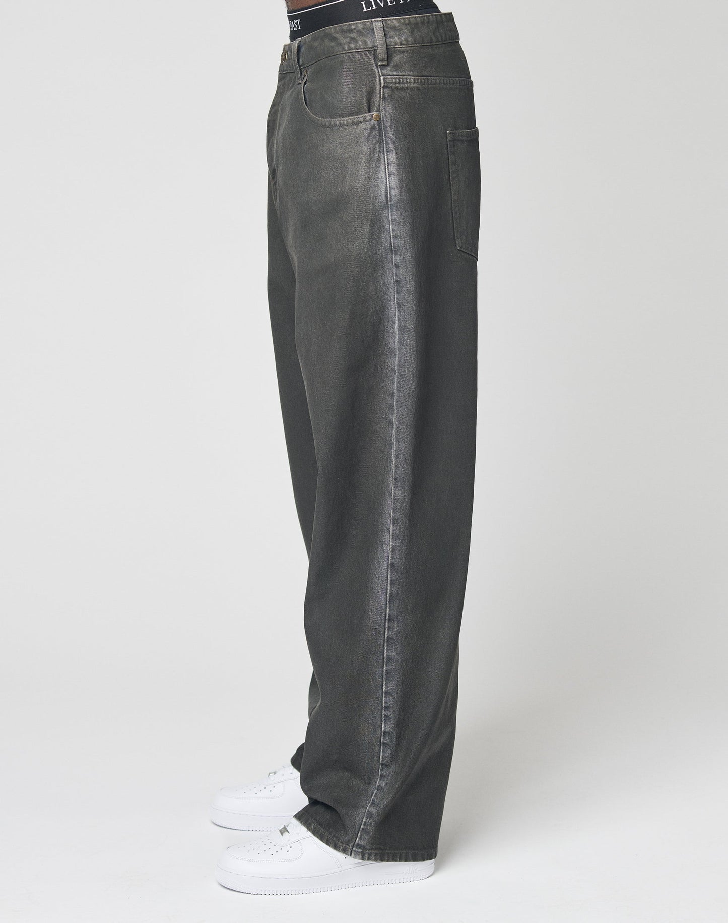 A person wearing LIVE FAST DIE YOUNG’s LFDY Coated Baggy Denim with straight legs and white sneakers stands against a plain white background, showing only the lower half of the body in side profile.