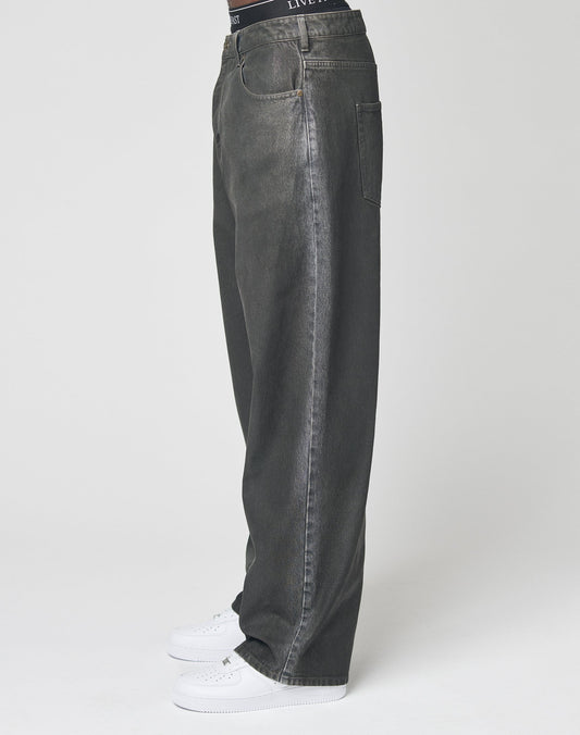 A person wearing LIVE FAST DIE YOUNG’s LFDY Coated Baggy Denim with straight legs and white sneakers stands against a plain white background, showing only the lower half of the body in side profile.