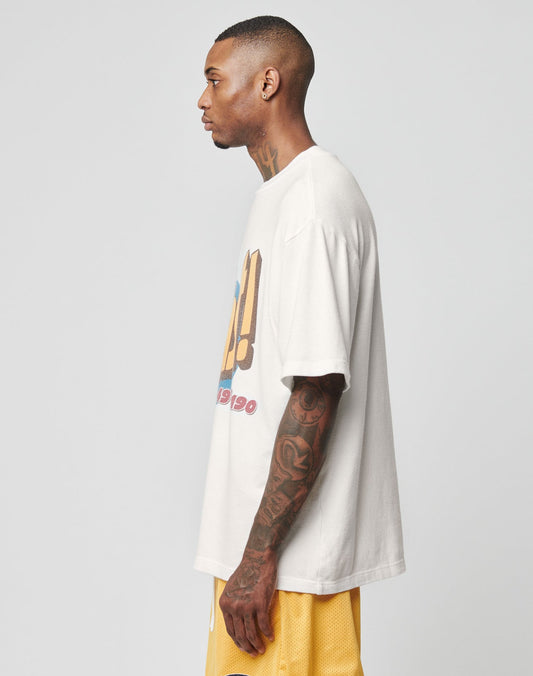 A man with short hair and visible tattoos stands in profile, wearing an oversize fit LFDY Adults Only Tee by LIVE FAST DIE YOUNG and yellow shorts, against a plain light gray background.