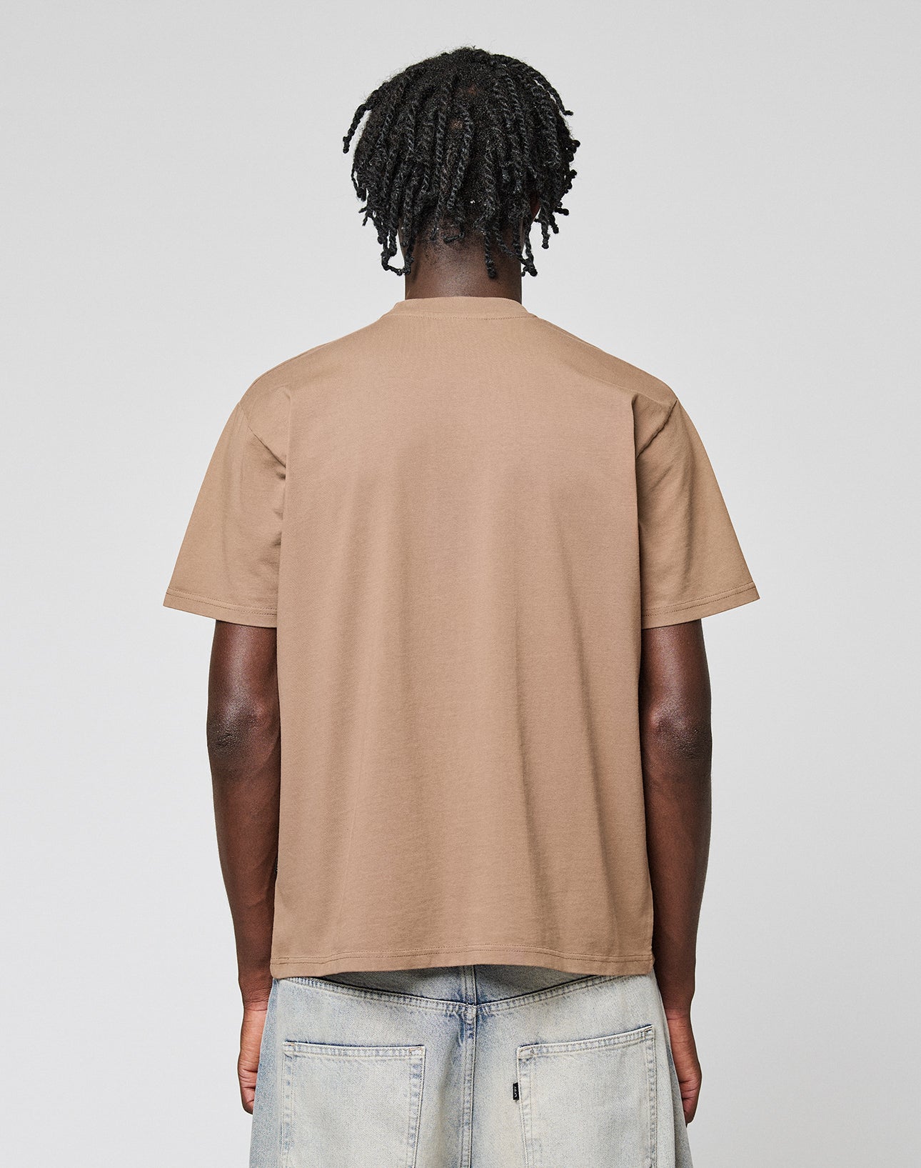 A person with short, twisted hair faces away from the camera, wearing the LIVE FAST DIE YOUNG Live Fast Plain Tee in light brown and light-wash denim jeans with large back pockets, standing against a plain light background.