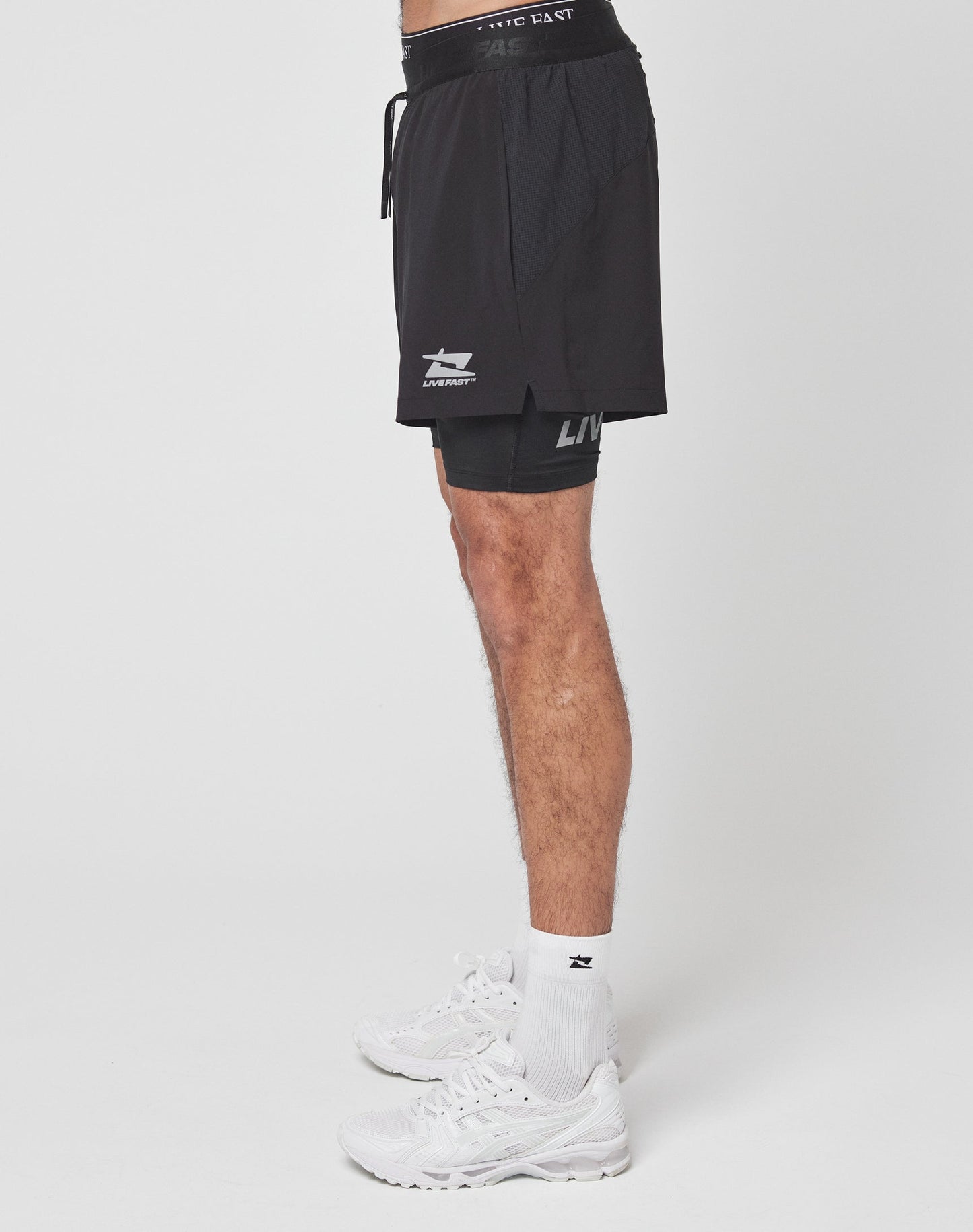 A close-up shows a person wearing black Live Fast Performance Running Shorts by LIVE FAST DIE YOUNG, featuring integrated tights and a prominent LIVE FAST logo on the left leg with part of their left and right thighs visible.
