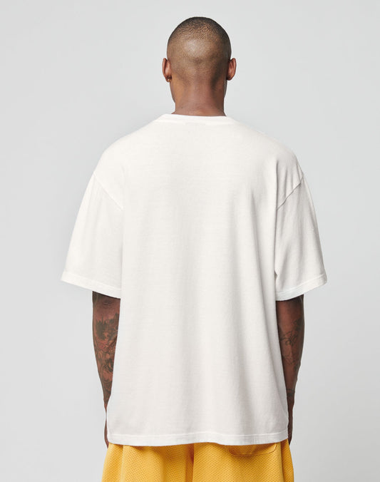 A person with closely cropped hair stands facing away, wearing an oversize fit plain white LFDY Adults Only Tee by LIVE FAST DIE YOUNG and yellow mesh shorts against a plain light gray background.