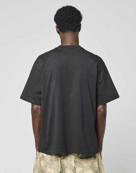 A person faces away from the camera wearing the LIVE FAST DIE YOUNG Live Fast Archive Tee in black with an oversize fit and light tie-dye shorts, set against a neutral gray background.