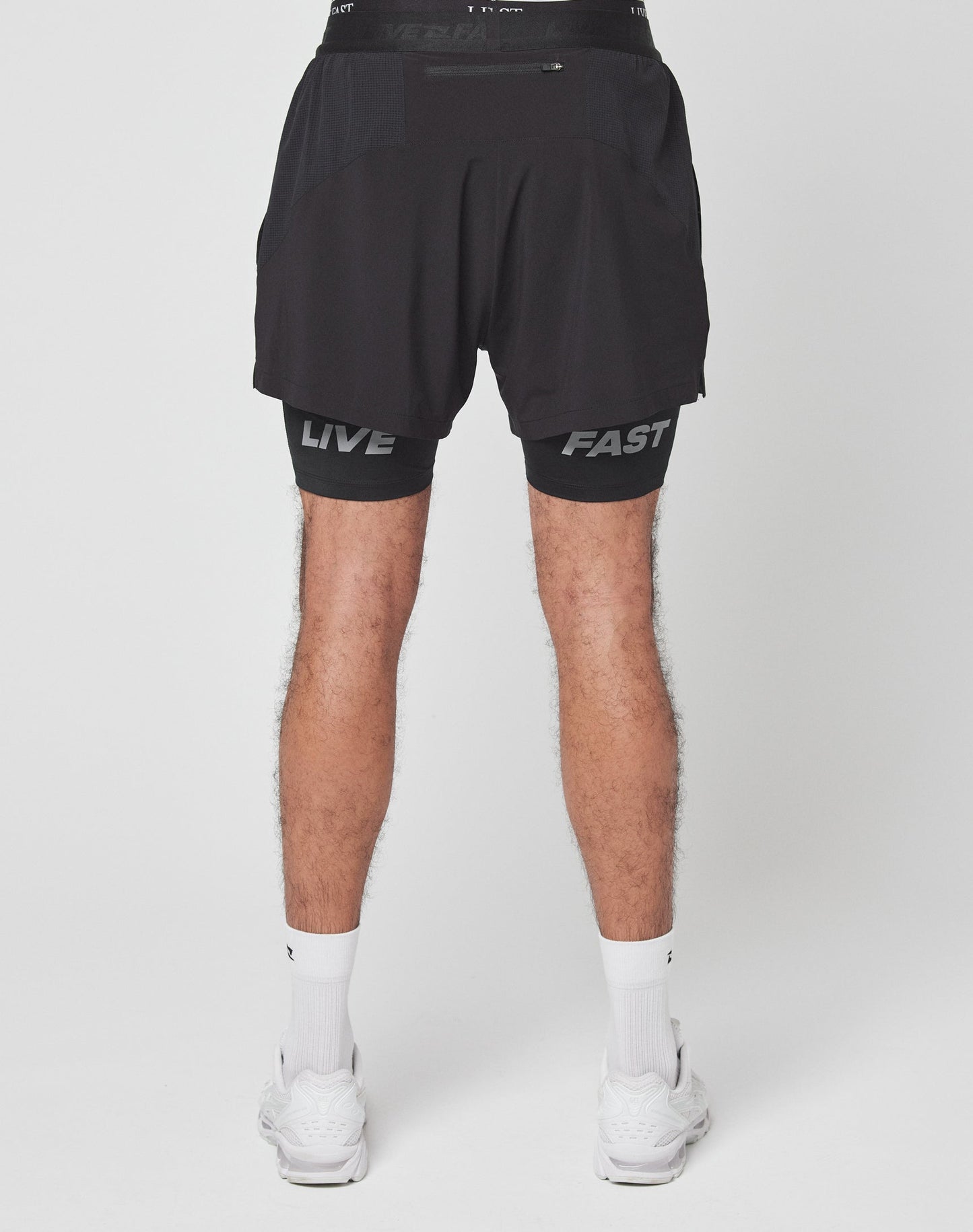 A person stands against a plain white background wearing LIVE FAST DIE YOUNG Live Fast Performance Running Shorts with integrierte Tights, a drawstring, and branded waistband, paired with white socks and sneakers.