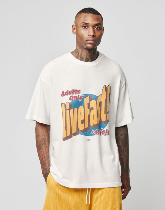 A tattooed man wears the LIVE FAST DIE YOUNG LFDY Adults Only Tee in white with bold lettering and yellow basketball shorts, standing against a plain light gray background.