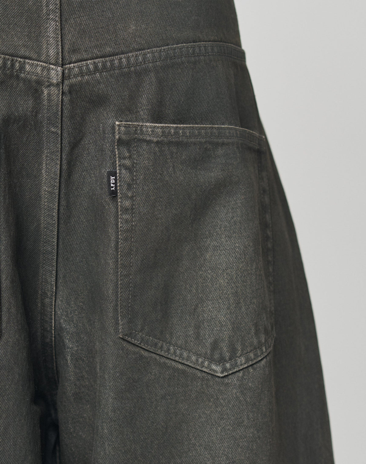 Close-up of the back pocket on LFDY Coated Baggy Denim by LIVE FAST DIE YOUNG, featuring dark gray 100% Baumwolle fabric, detailed stitching, a small black tag with white text, and a gerades Bein for a modern look.