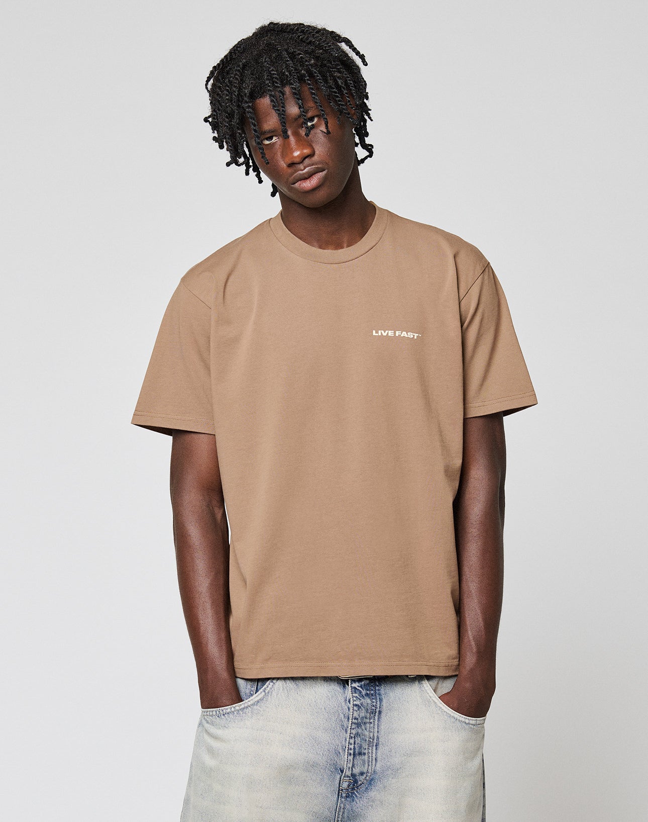 A young man with short dreadlocks wears the LIVE FAST DIE YOUNG Live Fast Plain Tee in brown (100% Baumwolle) featuring a subtle frontprint, paired with faded blue jeans. He stands against a light gray background, hands in his pockets.