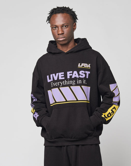 A person wears the LIVE FAST DIE YOUNG LFDY Racing Hooded black hoodie with glossy purple and yellow graphics that say LIVE FAST Everything in it, paired with matching black pants, against a plain light background.