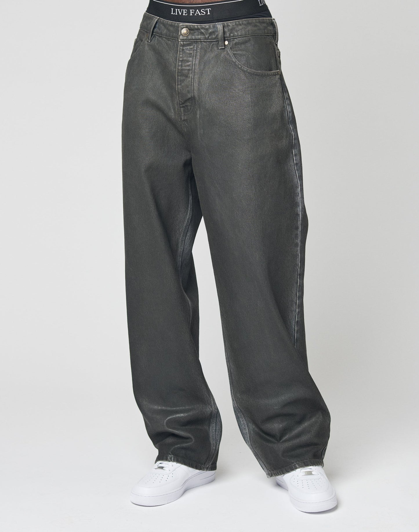 A person in LFDY Coated Baggy Denim by LIVE FAST DIE YOUNG, dark gray with a black waistband labeled LIVE FAST, and white sneakers stands against a plain white background. Their upper body is not visible.