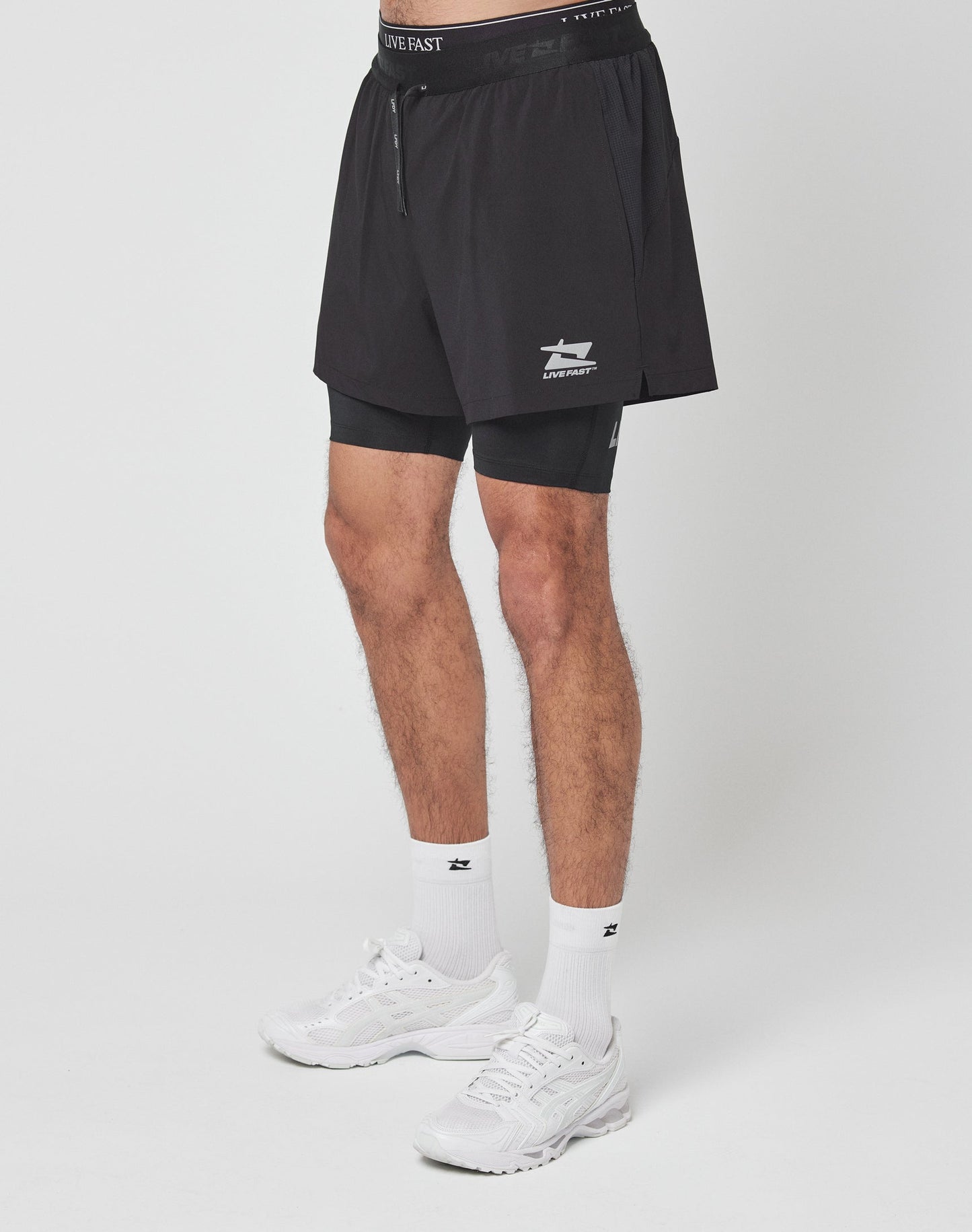 Someone wearing black Live Fast Performance Running Shorts by LIVE FAST DIE YOUNG places a smartphone in the side pocket, with a small zipper pocket and integrated tights details visible nearby.