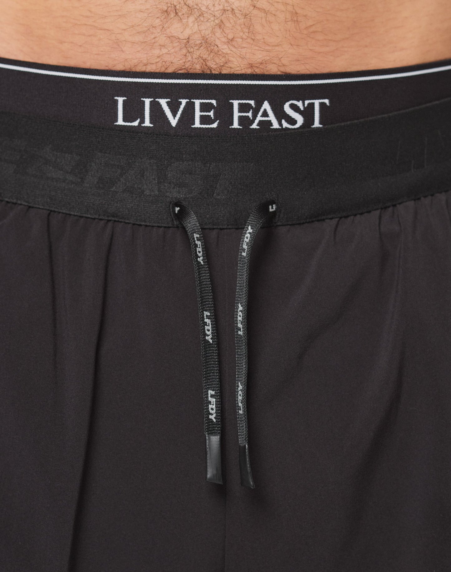 Close-up of a person’s lower torso wearing Live Fast Performance Running Shorts by LIVE FAST DIE YOUNG. The black shorts have an elastic waistband with white LIVE FAST branding and black drawstrings with white text in front.