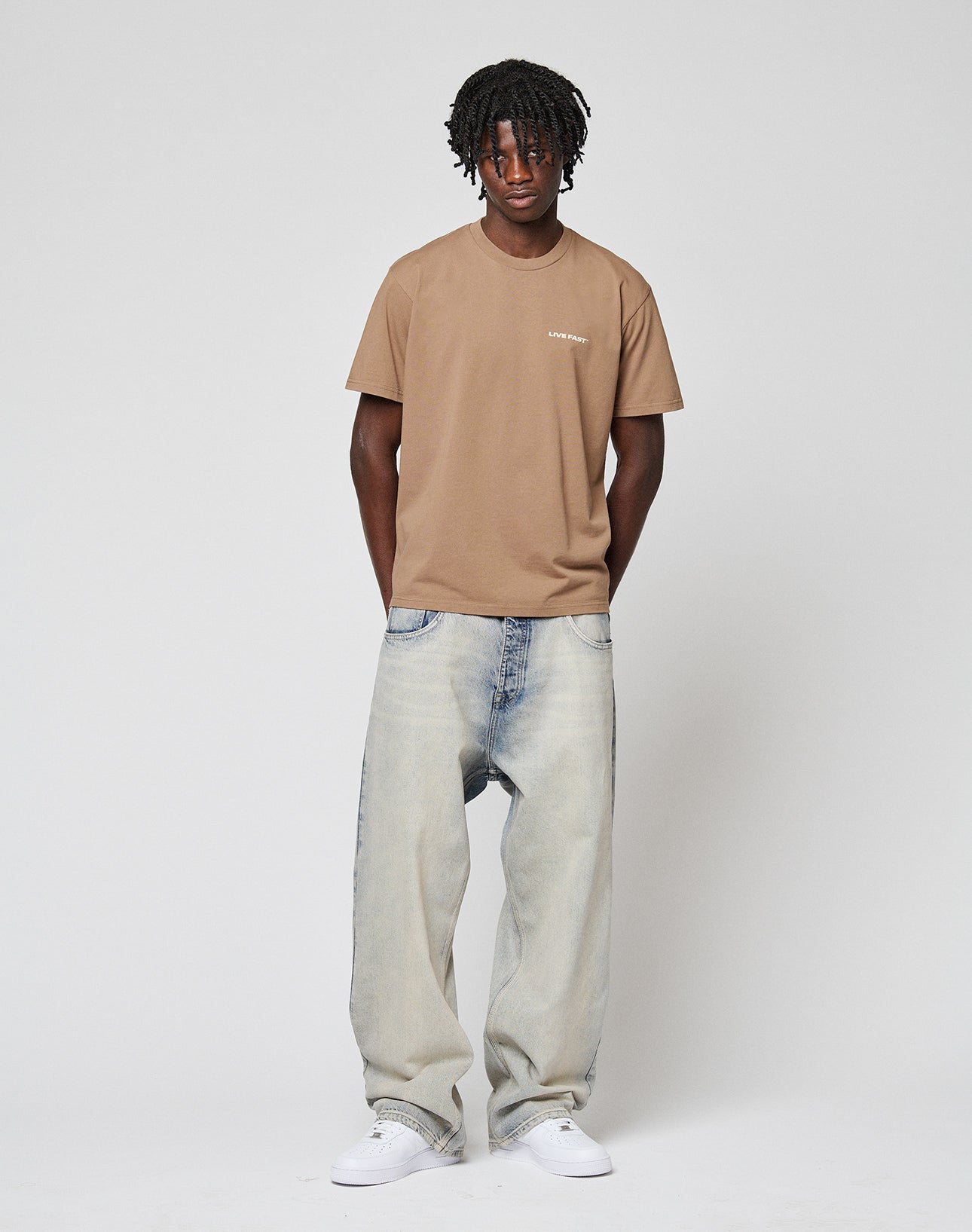 A person with shoulder-length locs stands against a plain background, wearing the LIVE FAST DIE YOUNG Live Fast Plain Tee (tan, 100% Baumwolle), light-wash baggy jeans, and white sneakers, with hands in their pockets.
