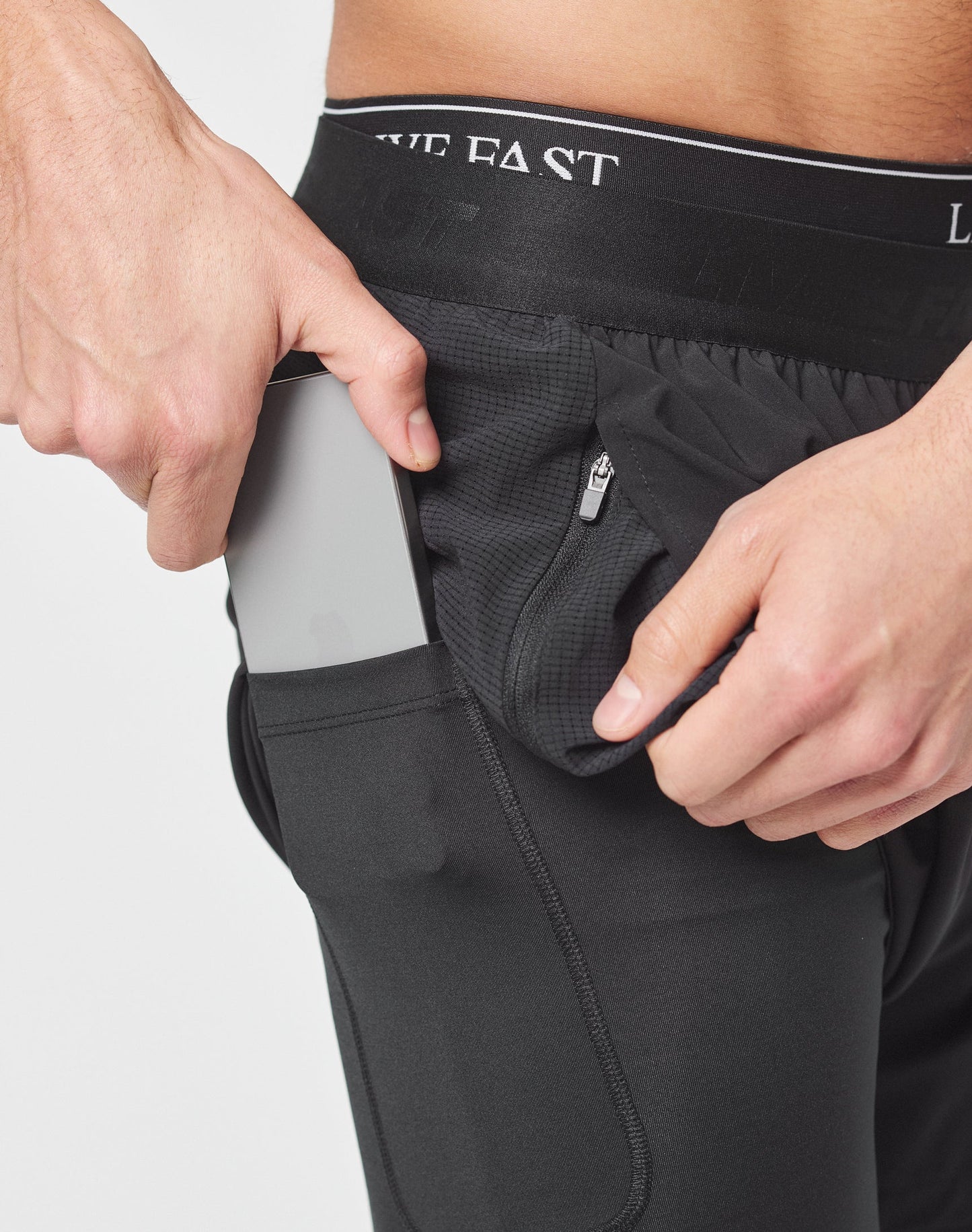 Someone wearing black Live Fast Performance Running Shorts by LIVE FAST DIE YOUNG places a smartphone in the side pocket, with a small zipper pocket and integrated tights details visible nearby.
