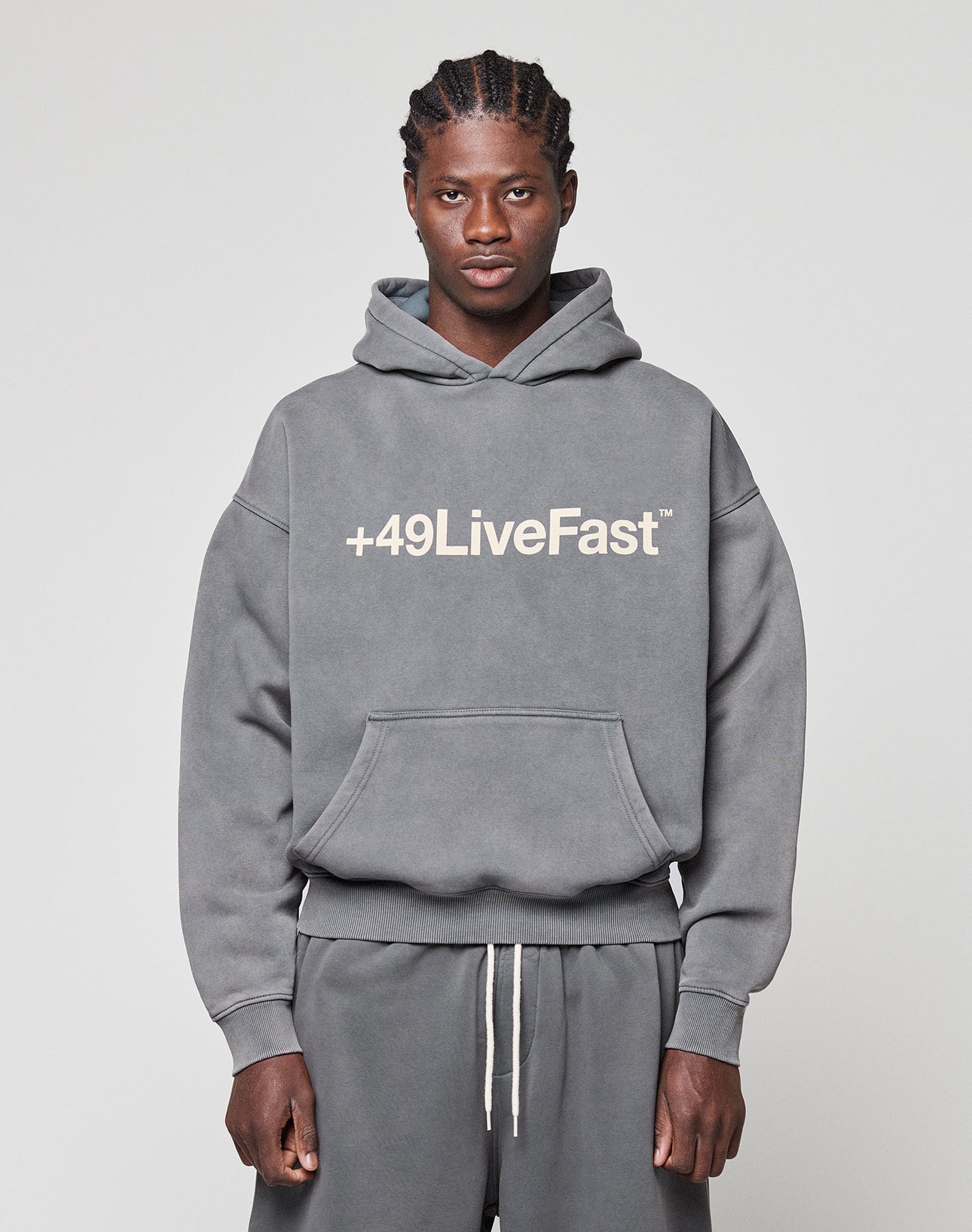 A man faces the camera in a gray LIVE FAST DIE YOUNG Basic +49 Hooded sweatshirt and matching sweatpants, featuring a bold white "+49LiveFast" front print. The background is plain and light-colored. | model