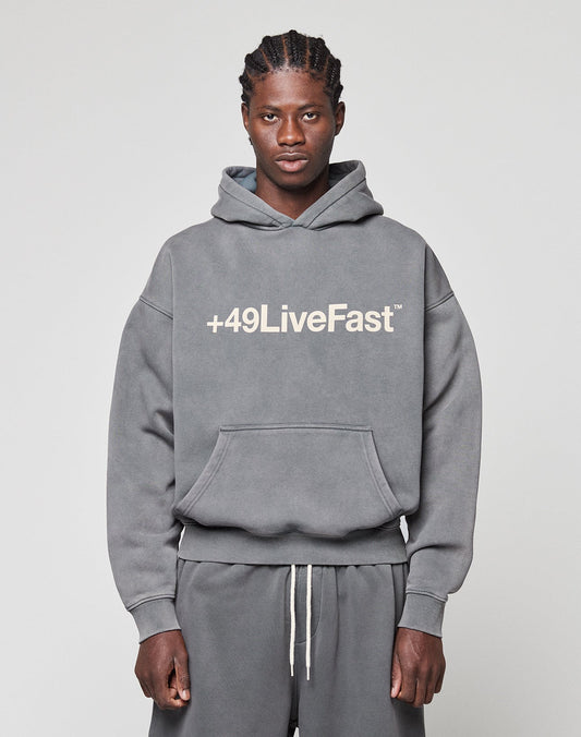 A man faces the camera in a gray LIVE FAST DIE YOUNG Basic +49 Hooded sweatshirt and matching sweatpants, featuring a bold white "+49LiveFast" front print. The background is plain and light-colored. | model