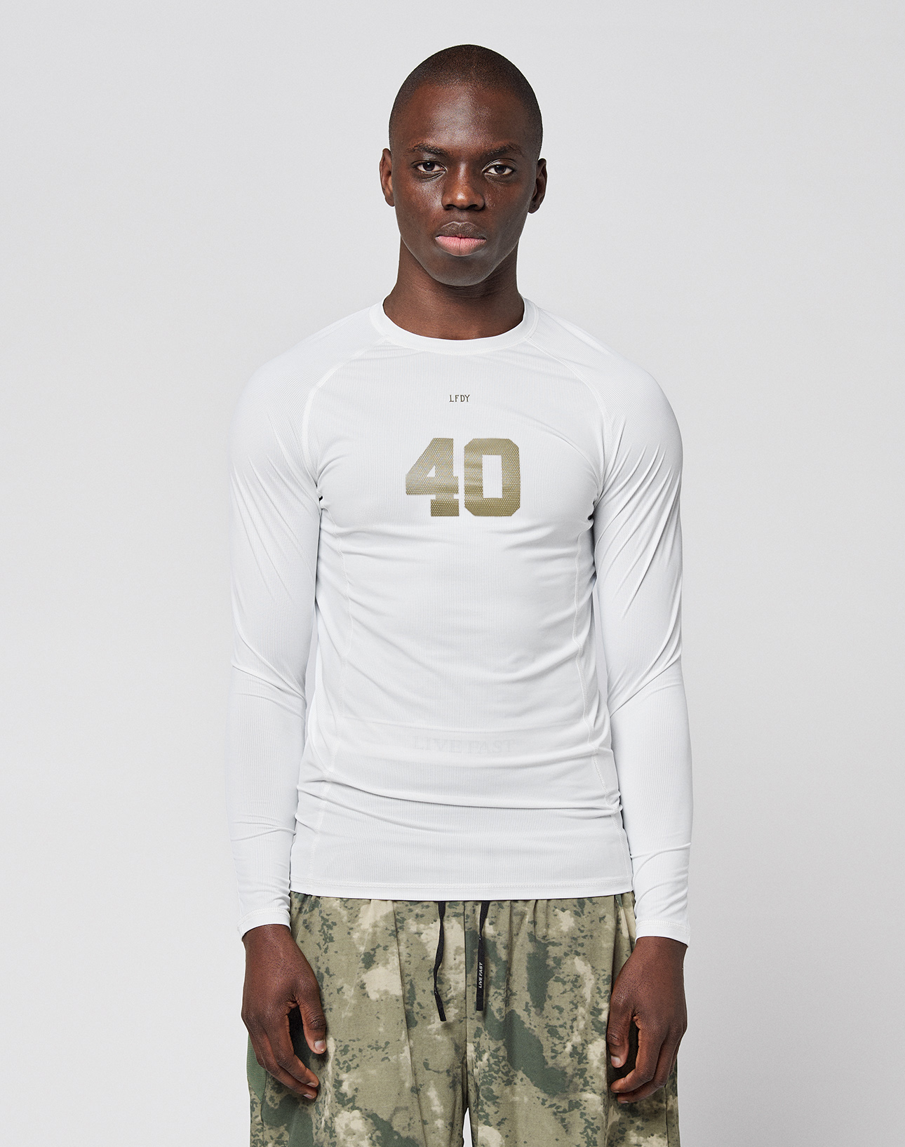 A person faces forward in a LIVE FAST DIE YOUNG LFDY Performance Slim Longsleeve, white with the number 40 on the front, and green-gray camouflage shorts made from leichtes Obermaterial, set against a plain light background. | model