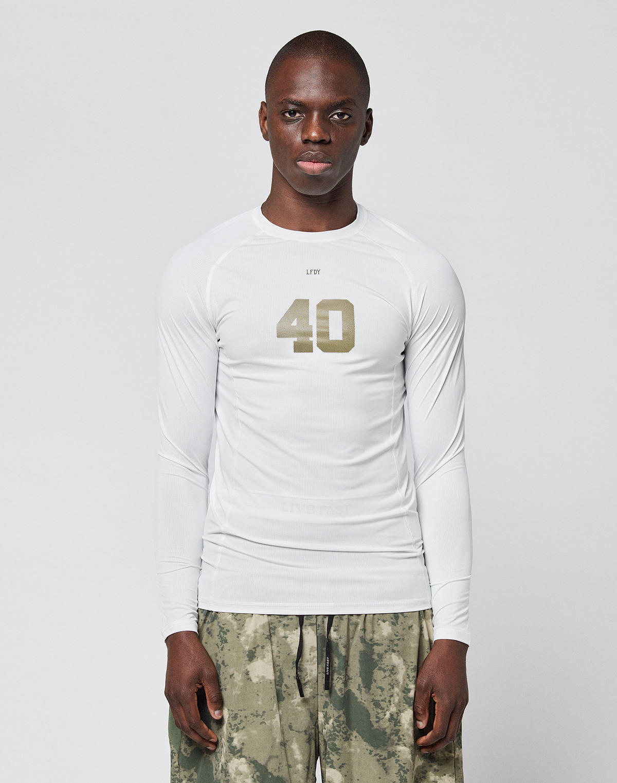 A person faces forward in a LIVE FAST DIE YOUNG LFDY Performance Slim Longsleeve, white with the number 40 on the front, and green-gray camouflage shorts made from leichtes Obermaterial, set against a plain light background. | model