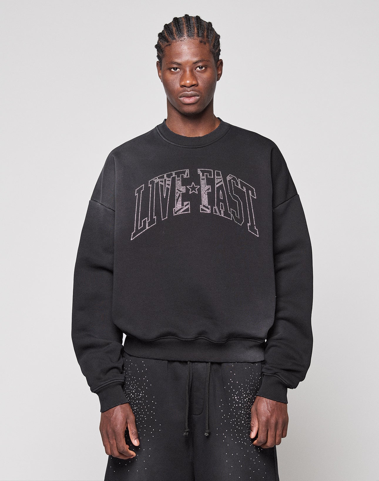 Against a plain background, a person wears the LIVE FAST DIE YOUNG Live Fast Rhinestone Sweater in an oversized fit with "LIVE FAST" on the front, paired with matching black pants featuring subtle white speckles.