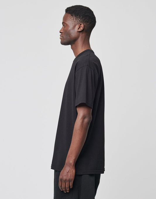 A man stands in profile facing left, wearing a regular fit black LFDY Blank Tee by LIVE FAST DIE YOUNG and dark pants against a plain light gray background.