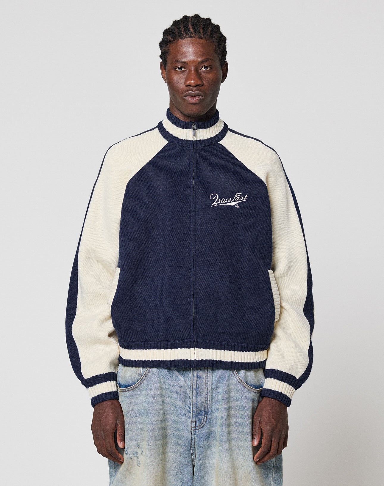 A man with braided hair wears a navy blue and cream Boxy Knit Zip Jacket by LIVE FAST DIE YOUNG, paired with light blue jeans, standing against a plain light background.
