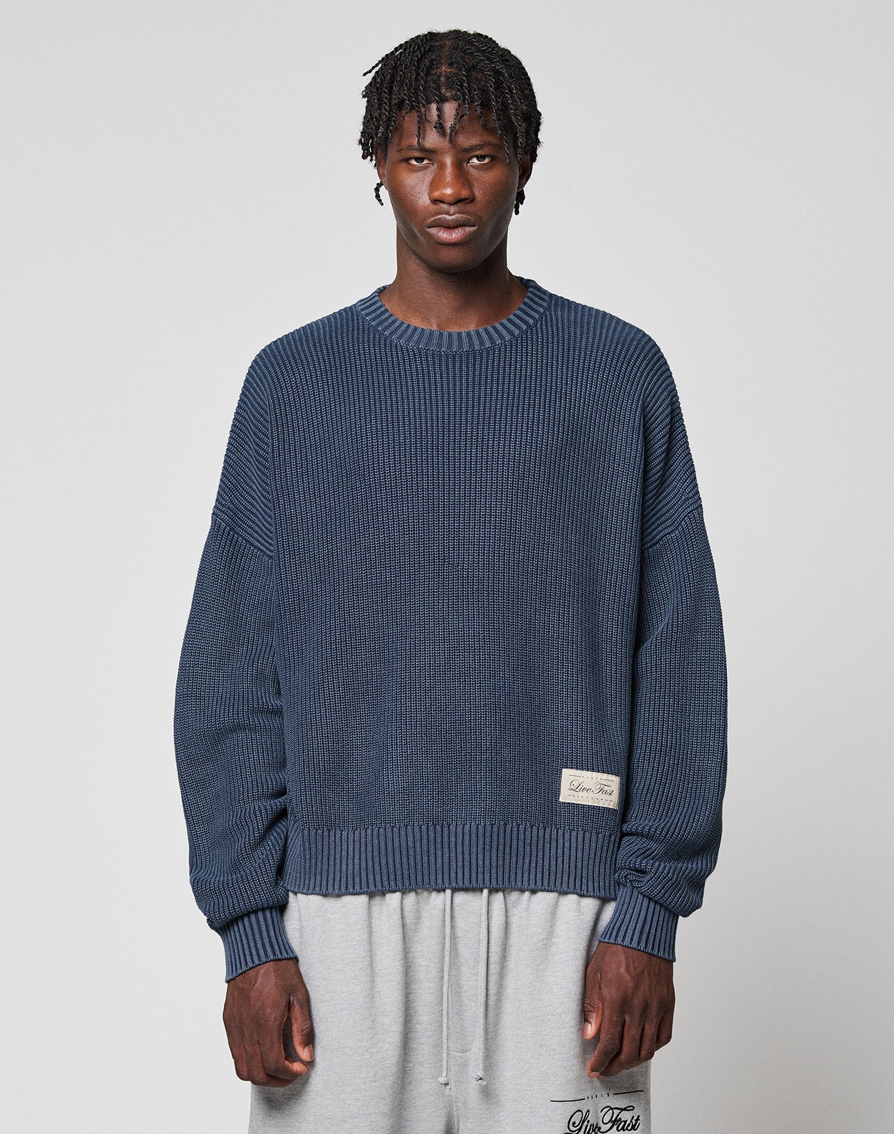 A man with short, twisted hair wears the LIVE FAST DIE YOUNG LFDY Knit Sweater in blue and light gray sweatpants, standing against a plain light gray background. | model