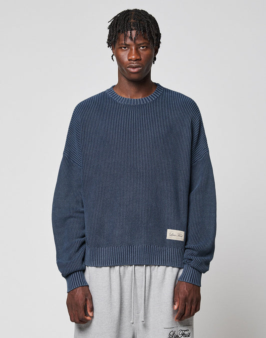 A man with short, twisted hair wears the LIVE FAST DIE YOUNG LFDY Knit Sweater in blue and light gray sweatpants, standing against a plain light gray background. | model
