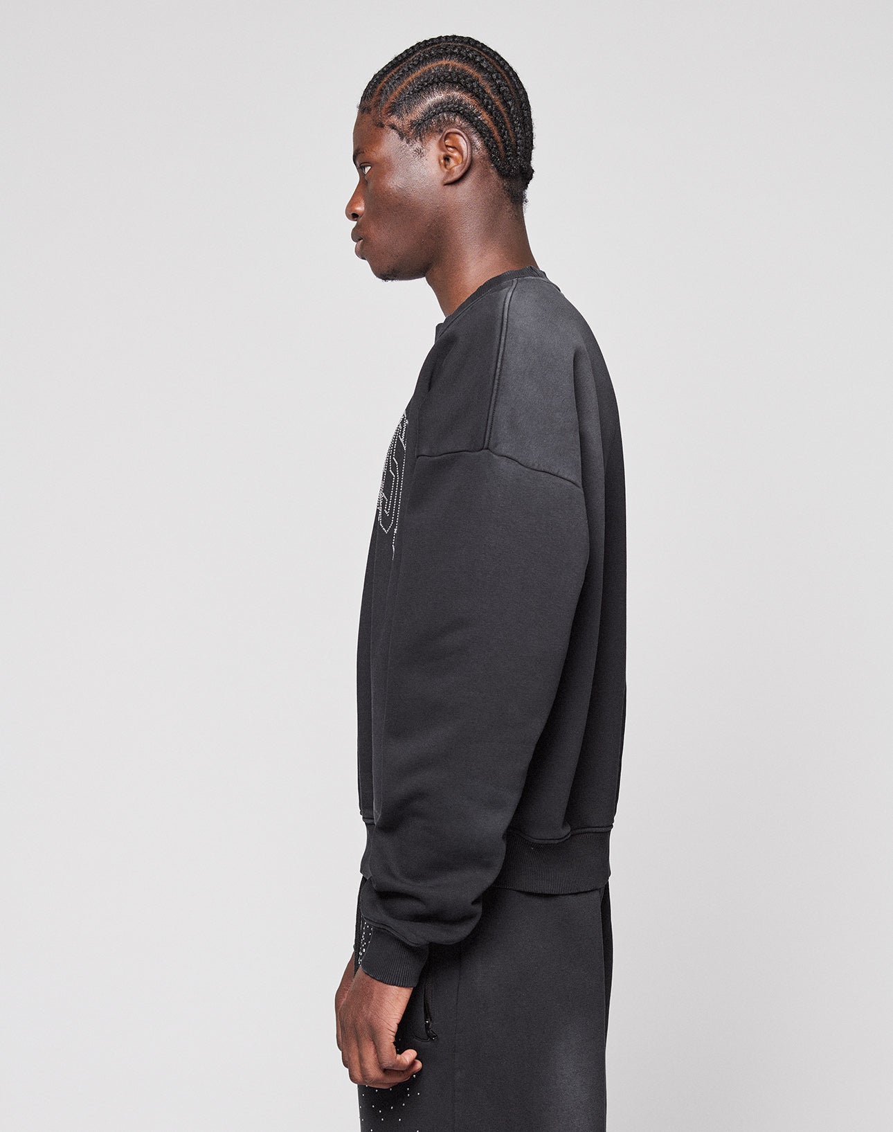 A person with braided hair stands in profile against a plain light gray background, wearing the LIVE FAST DIE YOUNG Live Fast Rhinestone Sweater in black and matching black pants.