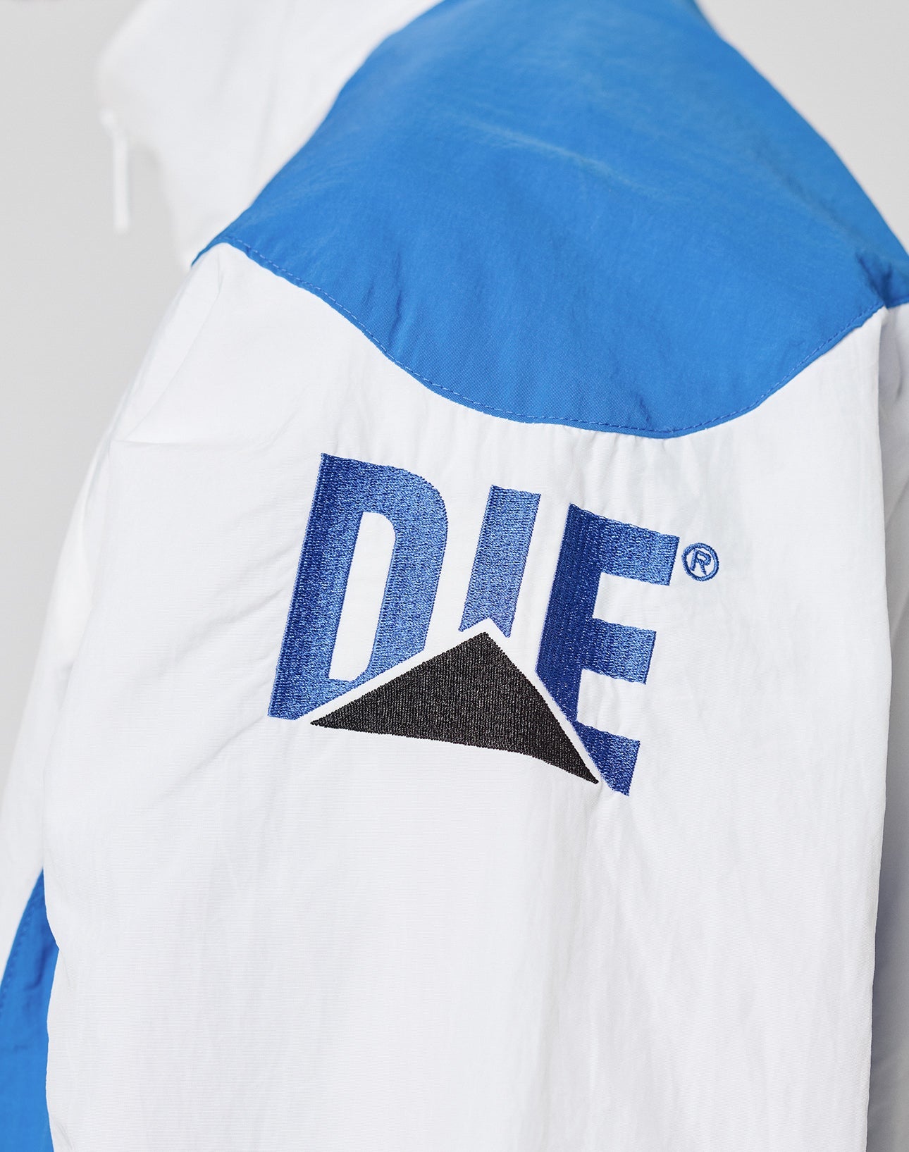 Close-up of the LFDY Trackjacket 3000 by LIVE FAST DIE YOUNG, a white and blue polyamide jacket with DIE in large blue letters and a black triangle on the back, plus a unique zip puller.