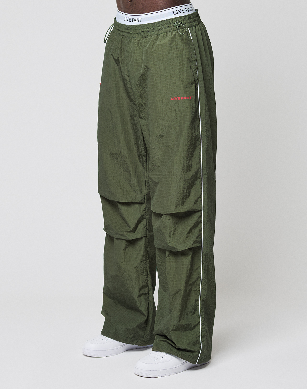 A person wears LFDY Nylon Trackpants by LIVE FAST DIE YOUNG—wide fit, olive green with white piping, LIVE FAST text on waistband and thigh, logo prints—paired with white sneakers, standing against a plain white background. | model