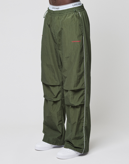 A person wears LFDY Nylon Trackpants by LIVE FAST DIE YOUNG—wide fit, olive green with white piping, LIVE FAST text on waistband and thigh, logo prints—paired with white sneakers, standing against a plain white background. | model