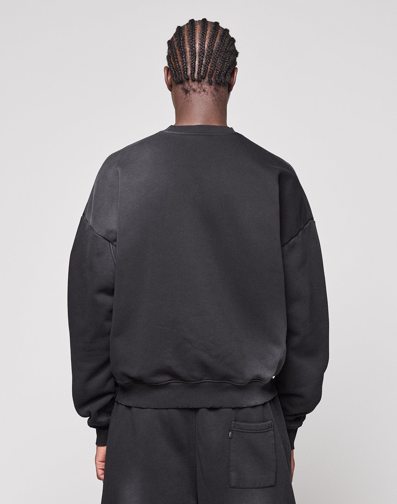 A person with braided hair is shown from the back, wearing the LIVE FAST DIE YOUNG Live Fast Rhinestone Sweater and black pants with a back pocket against a plain light background.