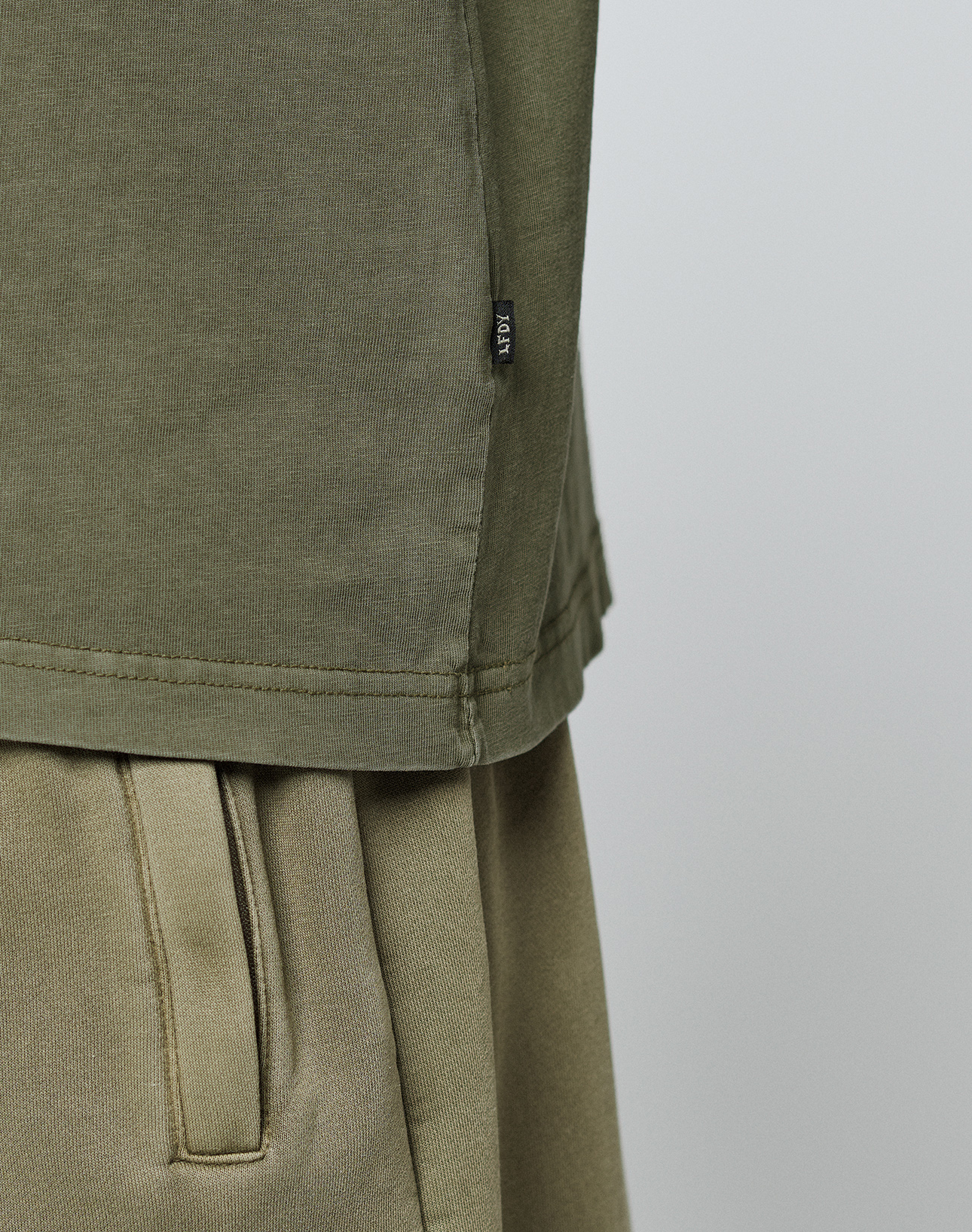 Close-up of a person wearing the olive green LIVE FAST DIE YOUNG Basic 210 Tee, made from 100% Baumwolle. The image shows only the midsection, highlighting the fabric texture and a small black tag on the side seam against a plain background.