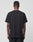 A person with short hair faces away from the camera, wearing a LIVE FAST DIE YOUNG LFDY Blank Tee—regular fit, plain black, short-sleeve, 100% cotton—and black pants against a plain light gray background.