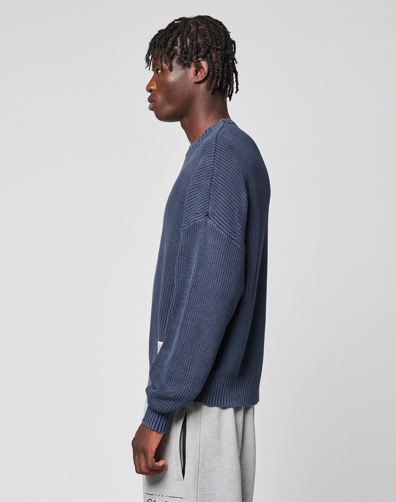 A young man with short dreadlocks stands in profile against a plain background, wearing the LIVE FAST DIE YOUNG LFDY Knit Sweater in an oversize fit and light gray sweatpants. The dropped shoulder design gives his look a relaxed edge.