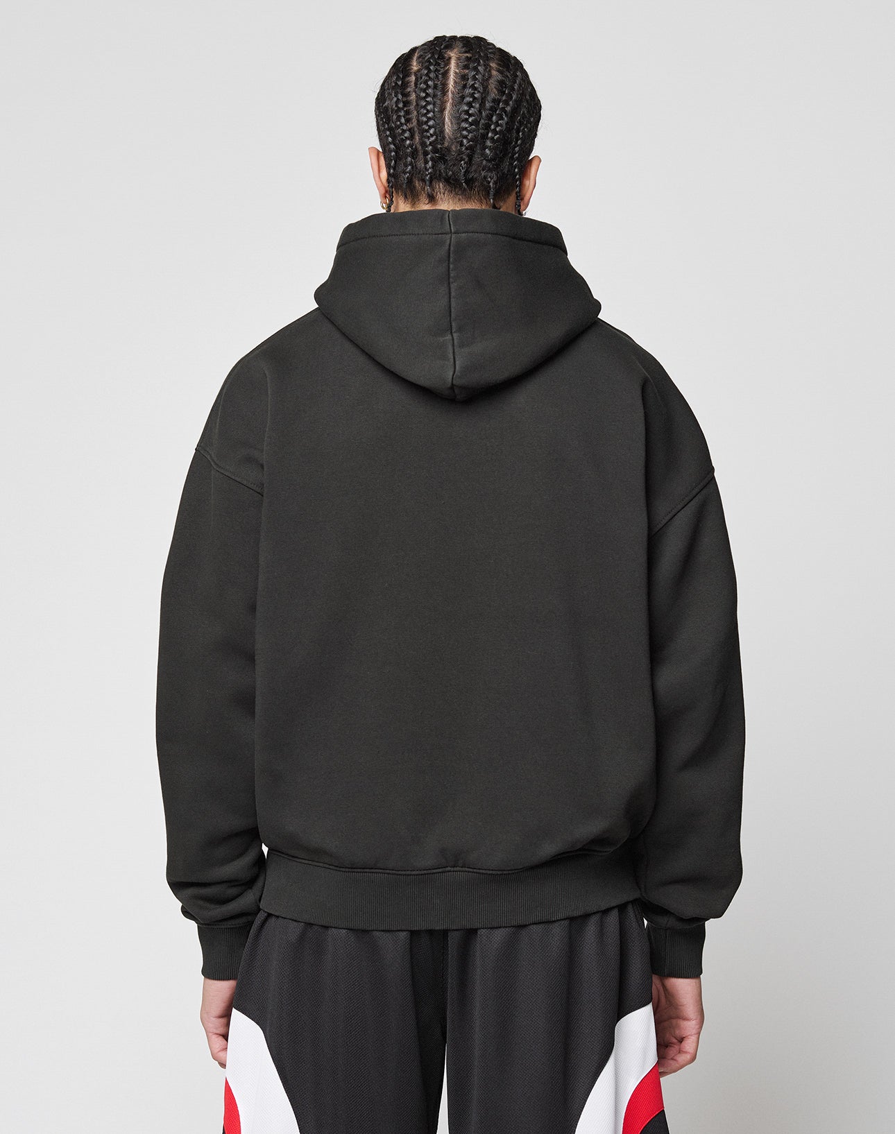 A person with braided hair is shown from the back, wearing the LIVE FAST DIE YOUNG Live Fast College Hooded oversize black hoodie (65% Baumwolle) and black pants with red and white accents, set against a plain light background.