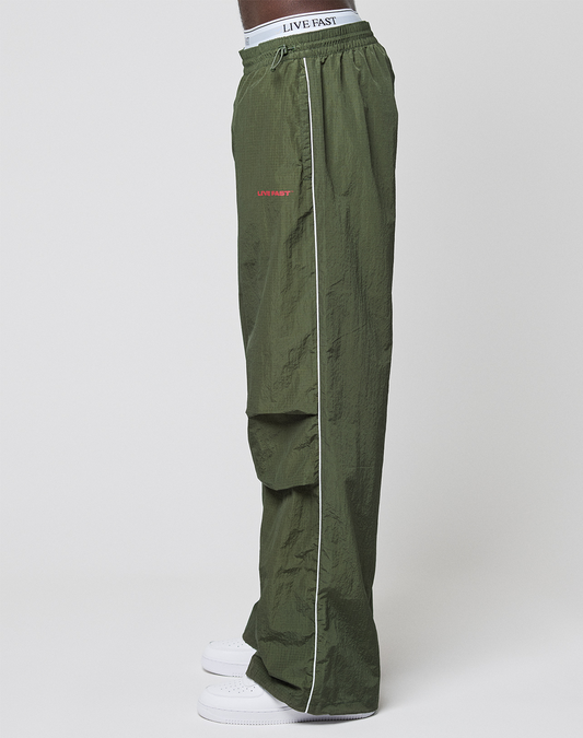 Someone stands sideways against a plain white background, wearing LFDY Nylon Trackpants by LIVE FAST DIE YOUNG in wide fit olive green with a white side stripe, LIVE FAST logo-print waistband, and white sneakers.