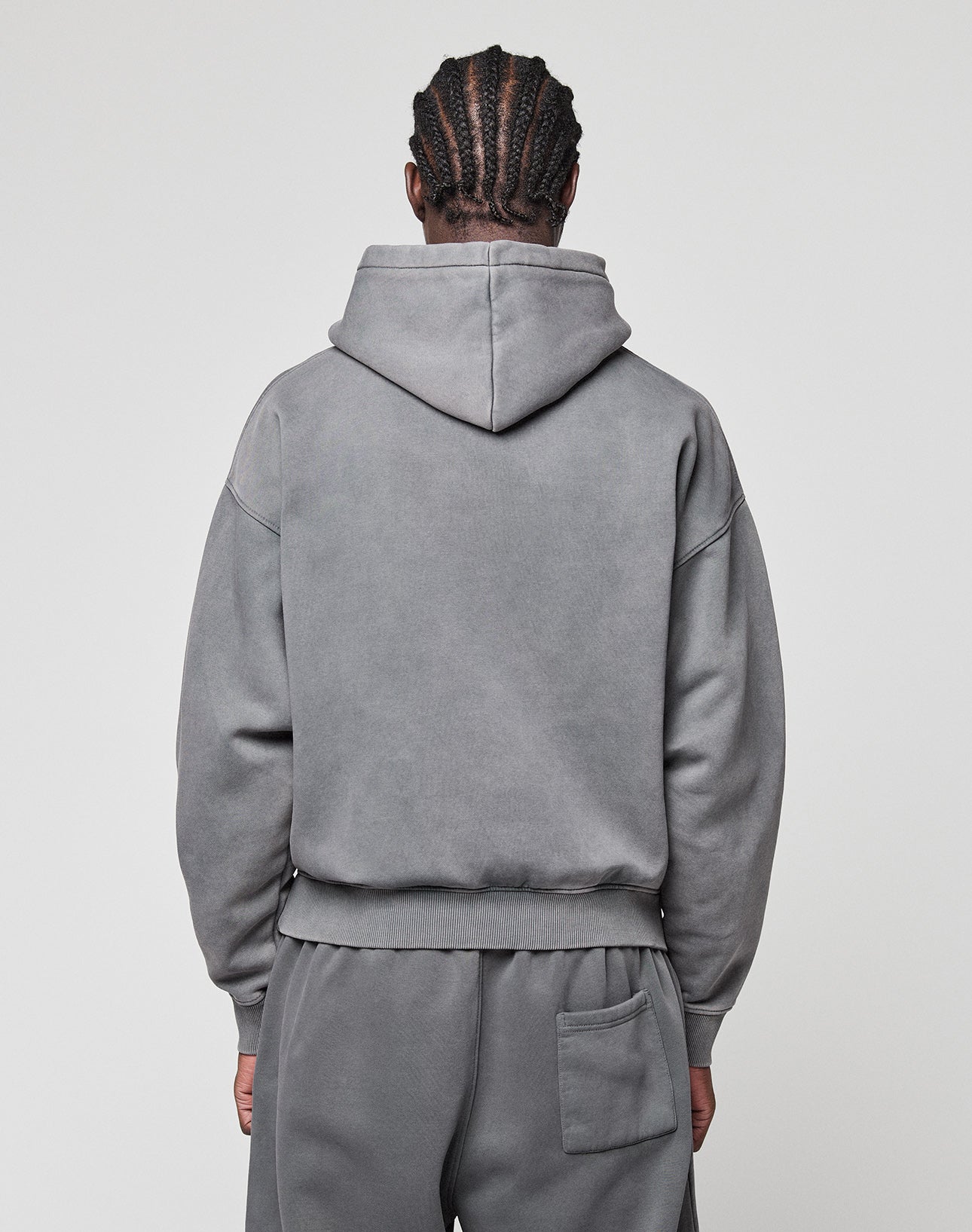 A person with braided hair is seen from behind wearing the LIVE FAST DIE YOUNG Basic +49 Hooded oversize grey hoodie and sweatpants set with a back pocket, made of Baumwolle, against a plain light background. The hood is down.