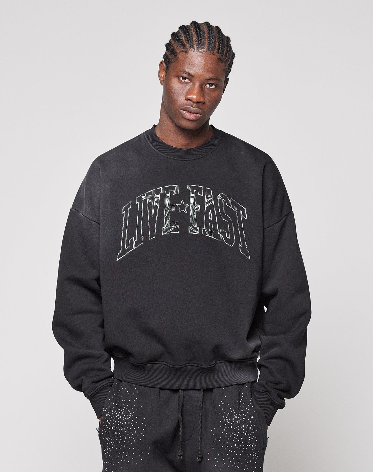 A man with braided hair wears the LIVE FAST DIE YOUNG Live Fast Rhinestone Sweater with matching black sweatpants featuring a white spray effect, standing against a plain light gray background.