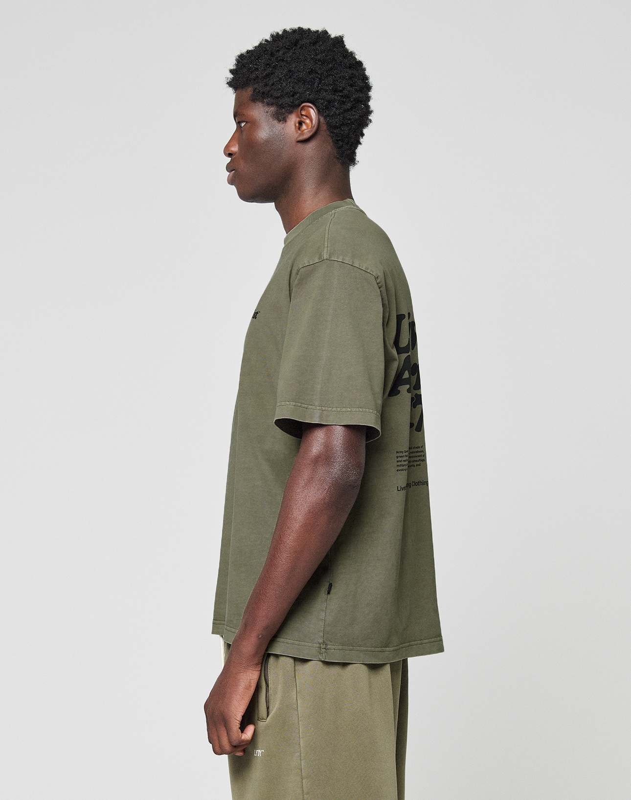 A person stands in profile against a plain background, wearing the LIVE FAST DIE YOUNG Basic 210 Tee in olive green, an oversize fit t-shirt made from 100% Baumwolle with black front and back print, paired with matching olive green shorts.