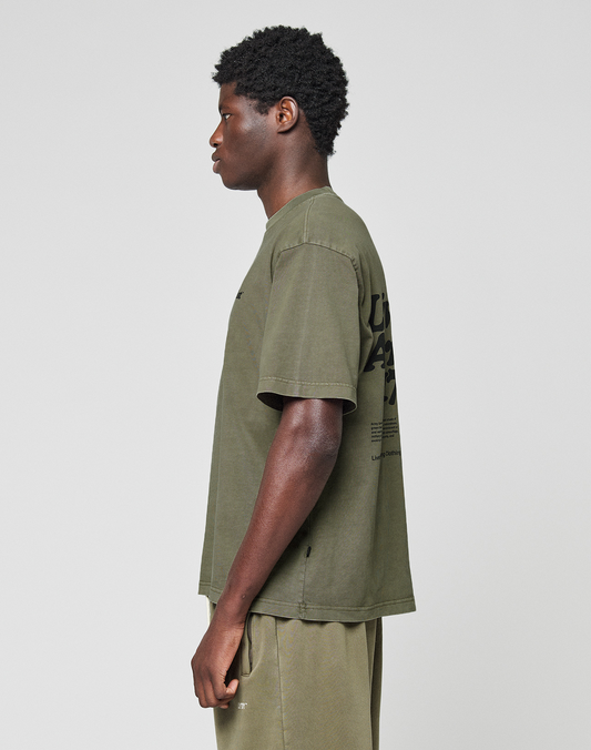 A person stands in profile against a plain background, wearing the LIVE FAST DIE YOUNG Basic 210 Tee in olive green, an oversize fit t-shirt made from 100% Baumwolle with black front and back print, paired with matching olive green shorts.