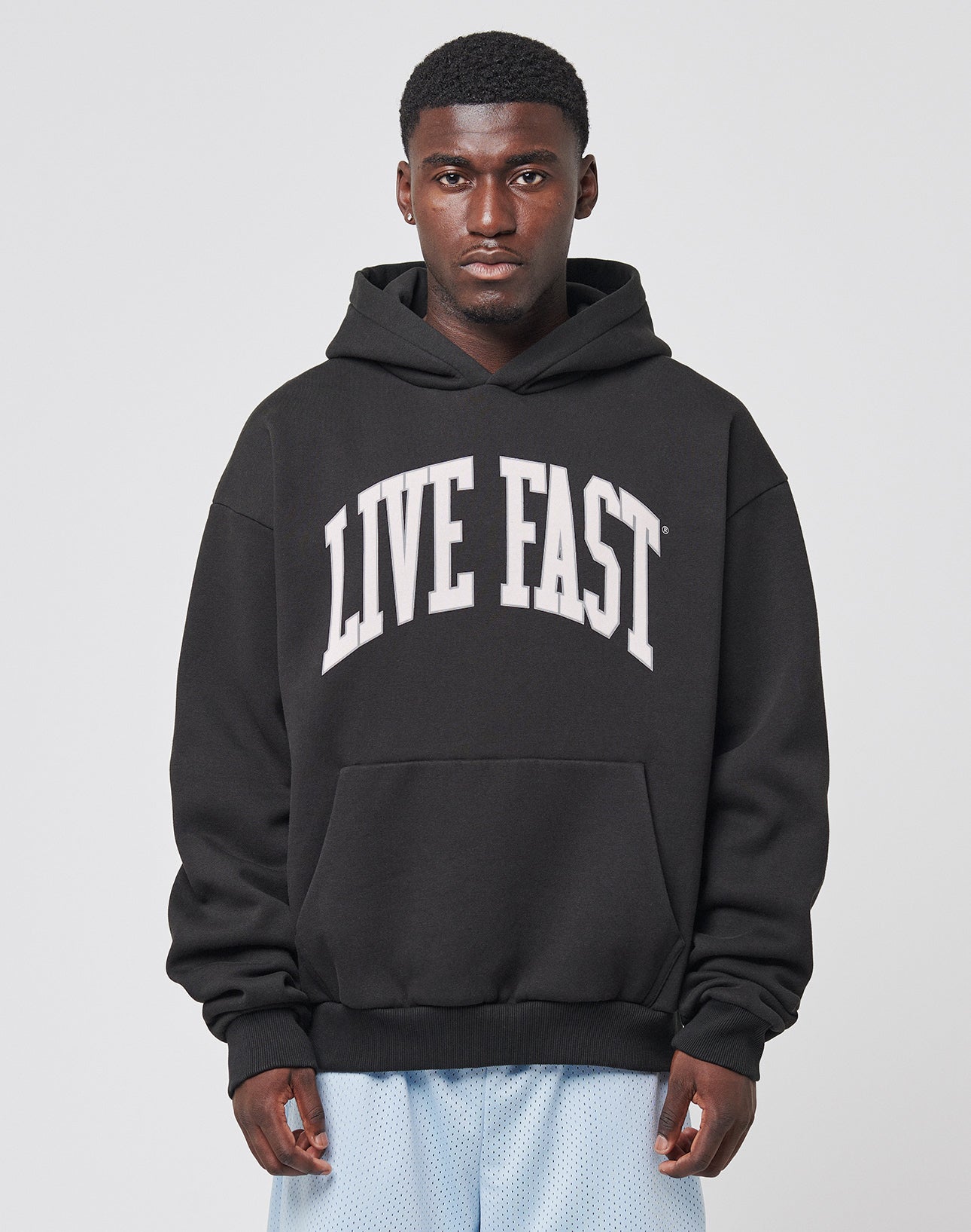 A man wearing the LIVE FAST DIE YOUNG Live Fast College Hooded, an oversize fit black cotton hoodie with bold frontprint, faces the camera against a plain light background.
