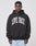 A man wearing the LIVE FAST DIE YOUNG Live Fast College Hooded, an oversize fit black cotton hoodie with bold frontprint, faces the camera against a plain light background.