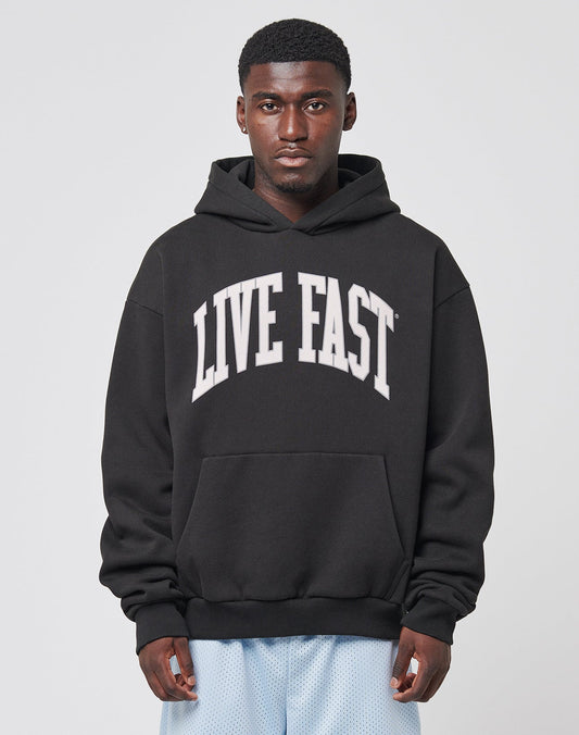 A man wearing the LIVE FAST DIE YOUNG Live Fast College Hooded, an oversize fit black cotton hoodie with bold frontprint, faces the camera against a plain light background.