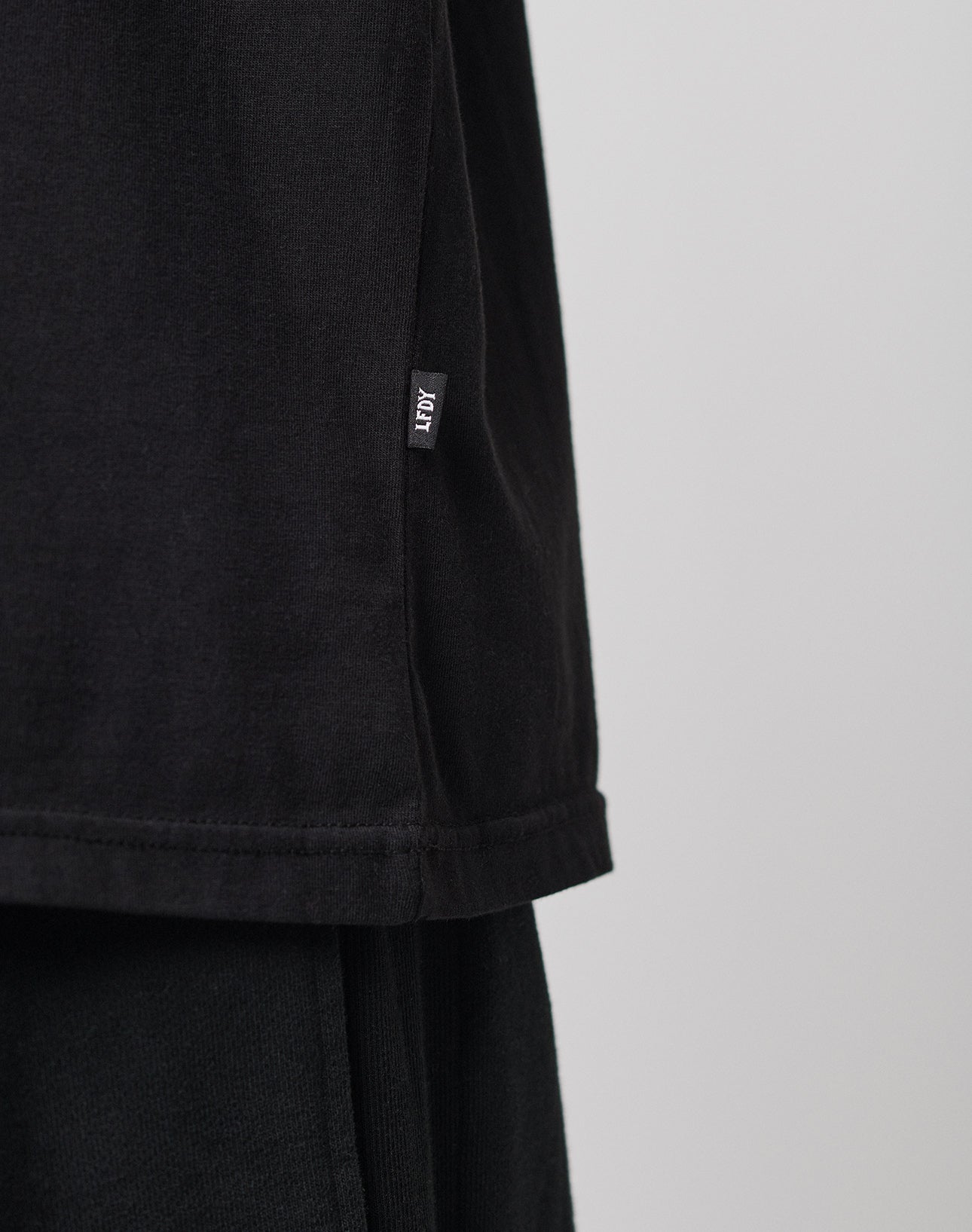 Close-up of the lower side of a black LIVE FAST DIE YOUNG LFDY Blank Tee with a small ADY tag sewn into the side seam, paired with black pants. Neutral, blurred background. Regular fit offers everyday comfort.